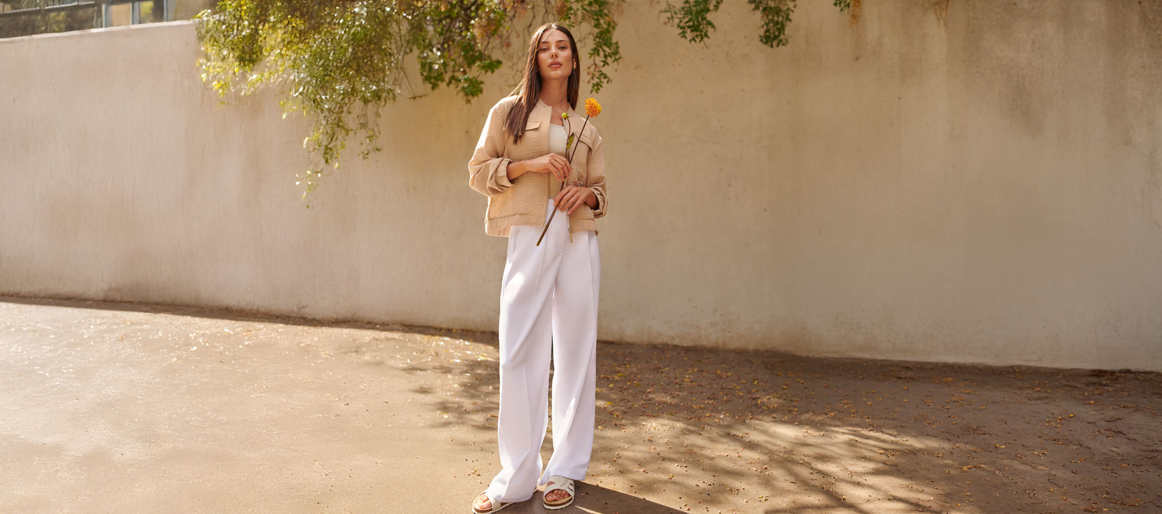 Donna con giacca beige, pantaloni bianchi e sandali, che tiene un fiore giallo.