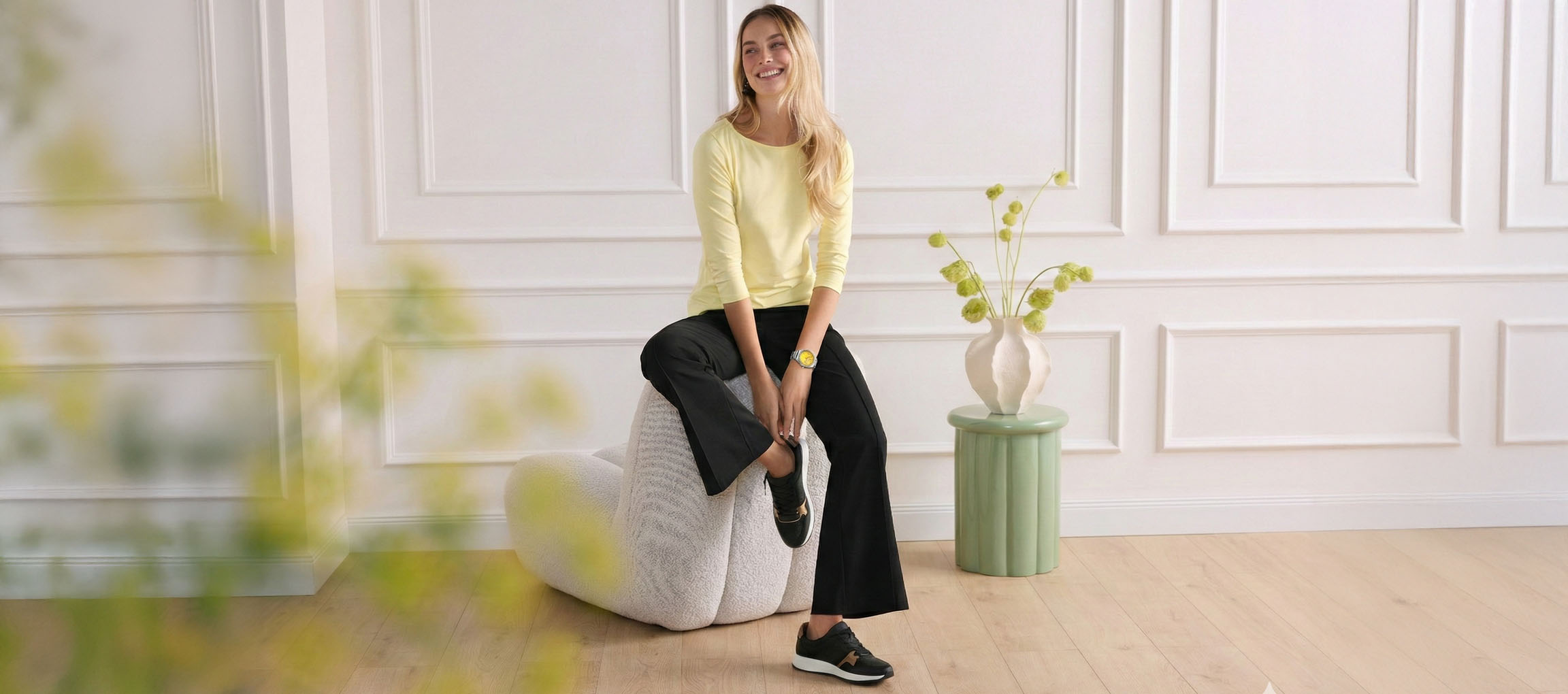 Femme en haut jaune et pantalon noir assise sur un pouf, souriante.