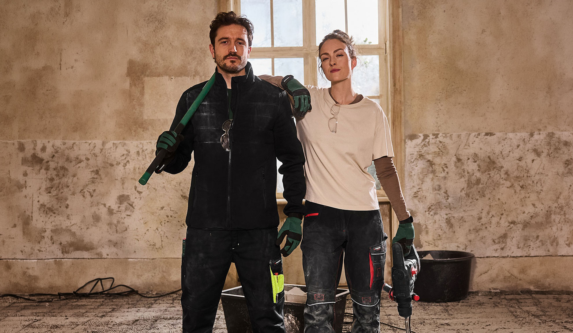 Deux ouvriers du bâtiment en tenue de travail, homme avec pelle et femme avec perceuse.