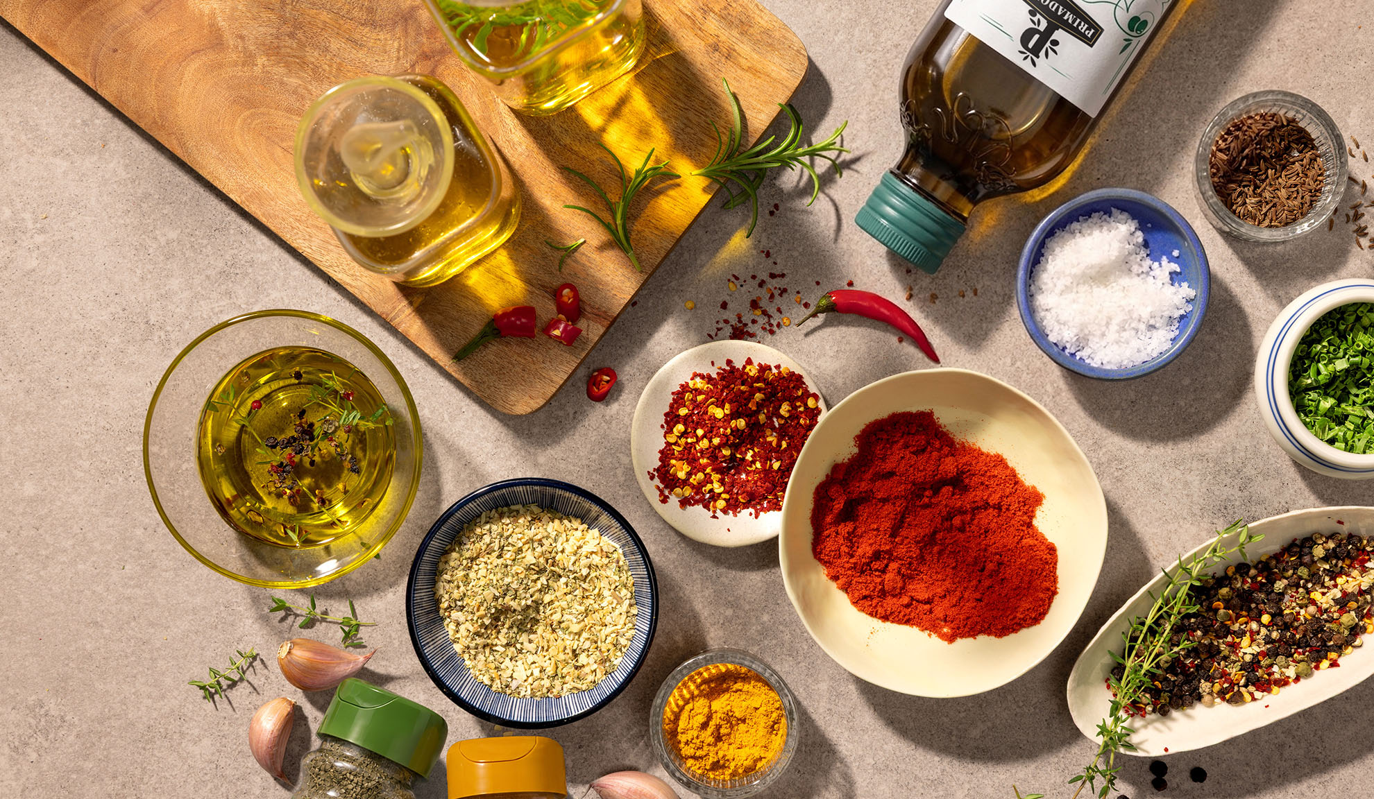 Olio d'oliva, spezie e peperoncini su un tagliere di legno e un piano di lavoro in cemento.