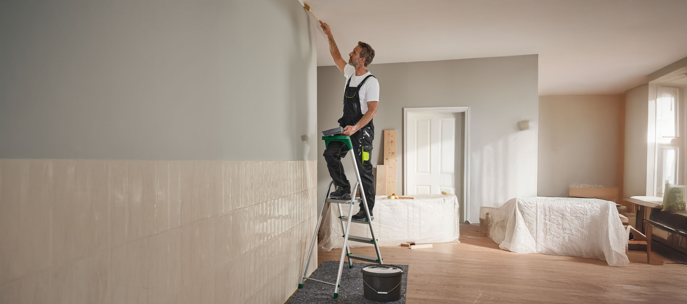 Ein Mann streicht mit einem Pinsel eine Wand und steht dabei auf einer Trittleiter.