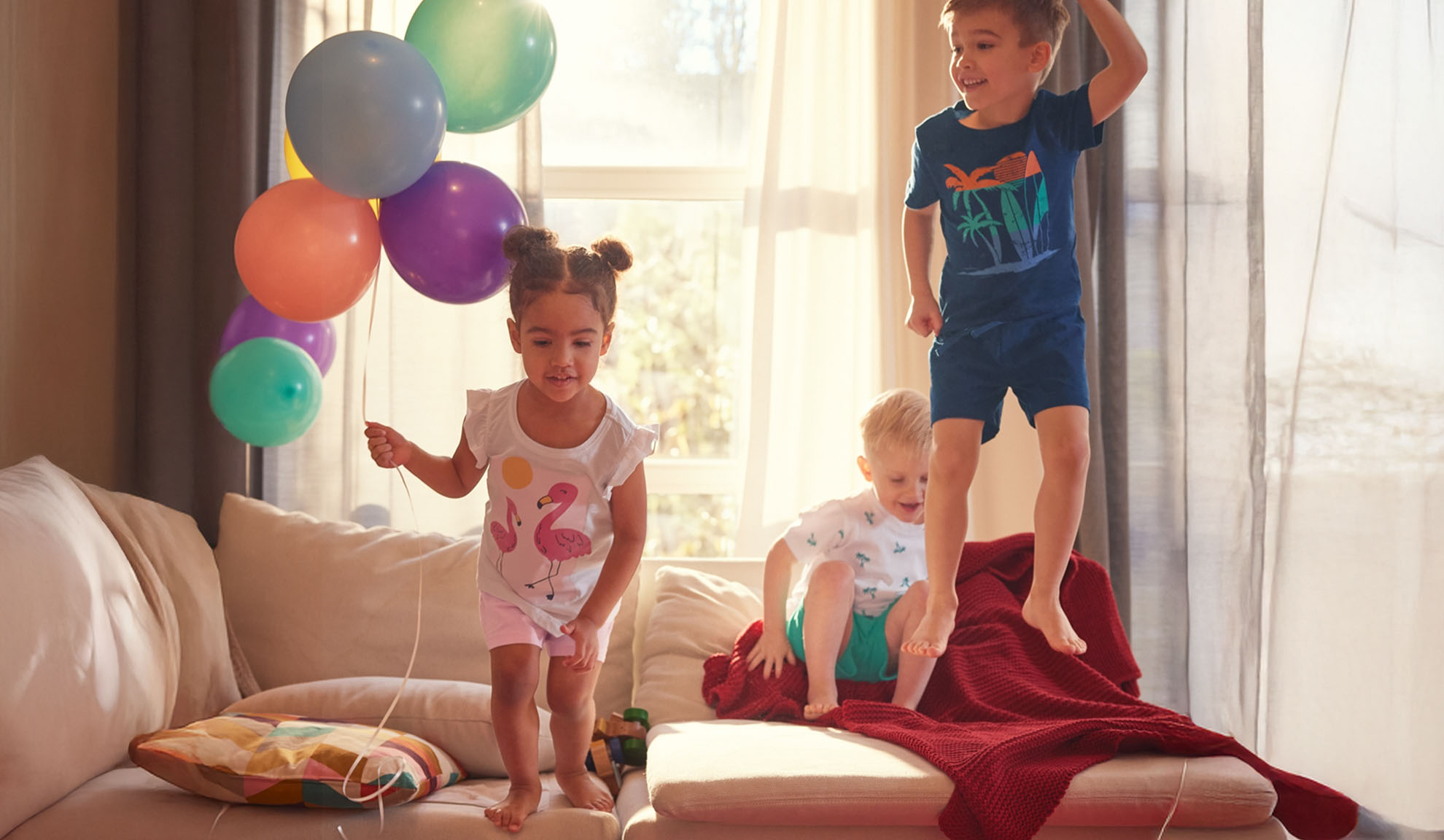 Trois enfants sautent joyeusement sur un canapé avec des ballons colorés.