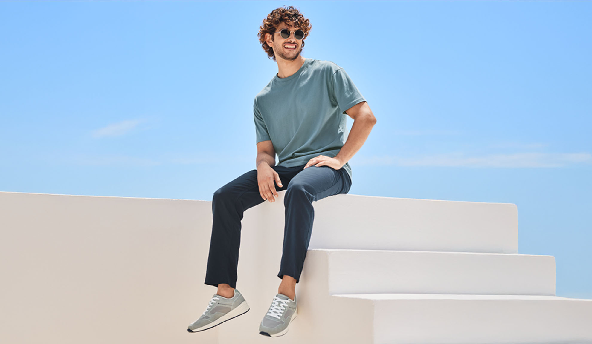 Mann in einem blauen T-Shirt und dunklen Hosen sitzt entspannt auf einer weißen Treppe.