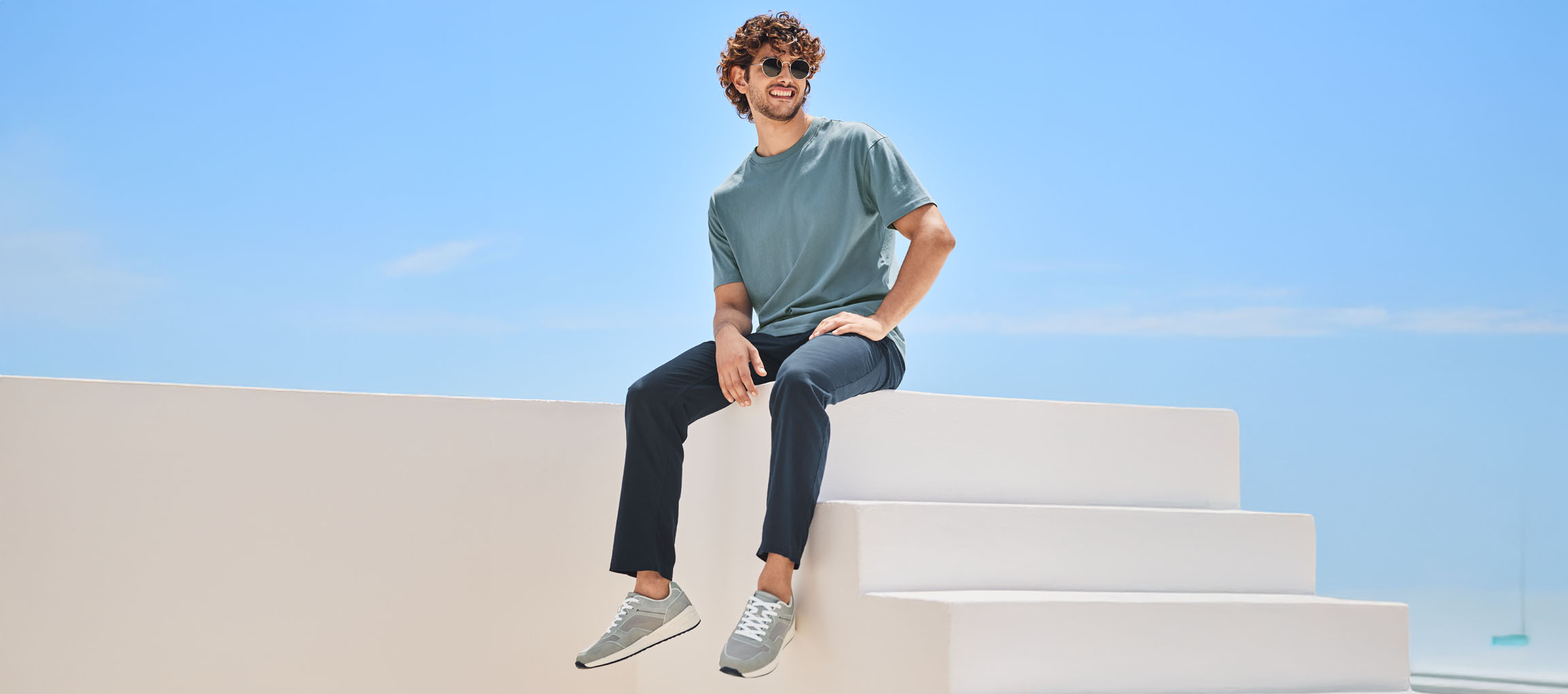 Mann in lässigem Outfit mit T-Shirt, Hose und New Balance Schuhen, sitzt auf einer weißen Treppe.