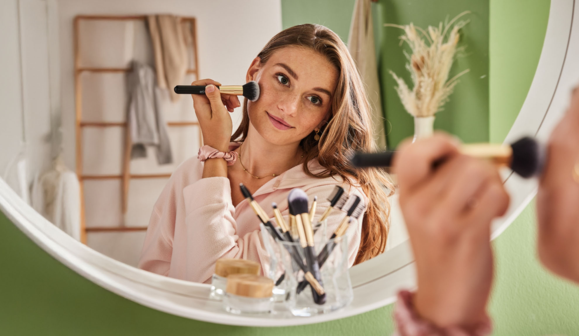 Frau trägt Make-up mit einem Pinsel auf, im ovalen Spiegel reflektiert.