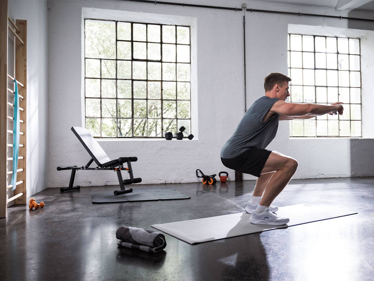 Homme faisant des squats dans une salle de sport avec un banc de musculation et des tapis.