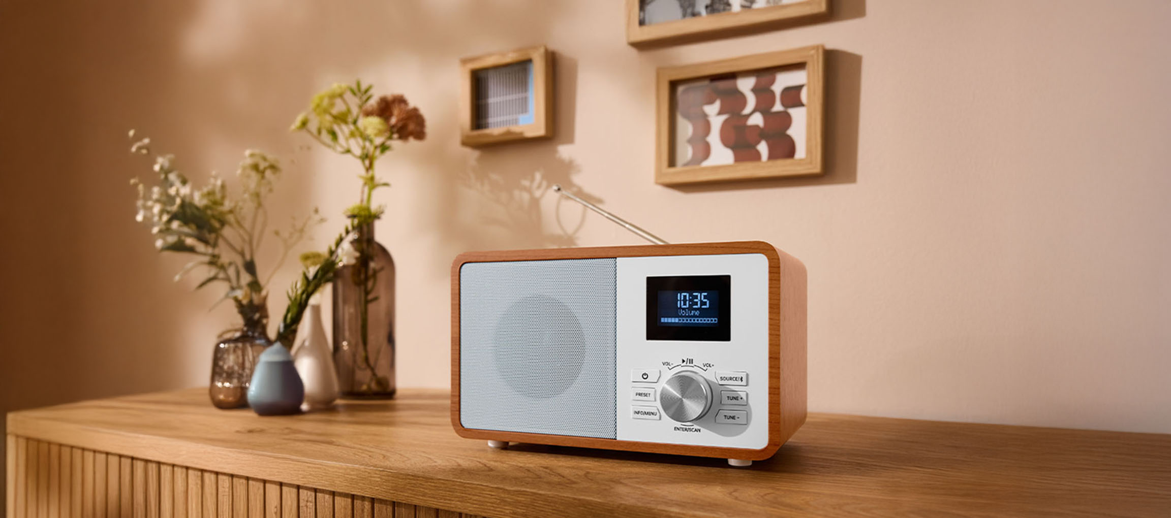 Una radio digitale in legno su una cassettiera, con vasi e quadri sulla parete.