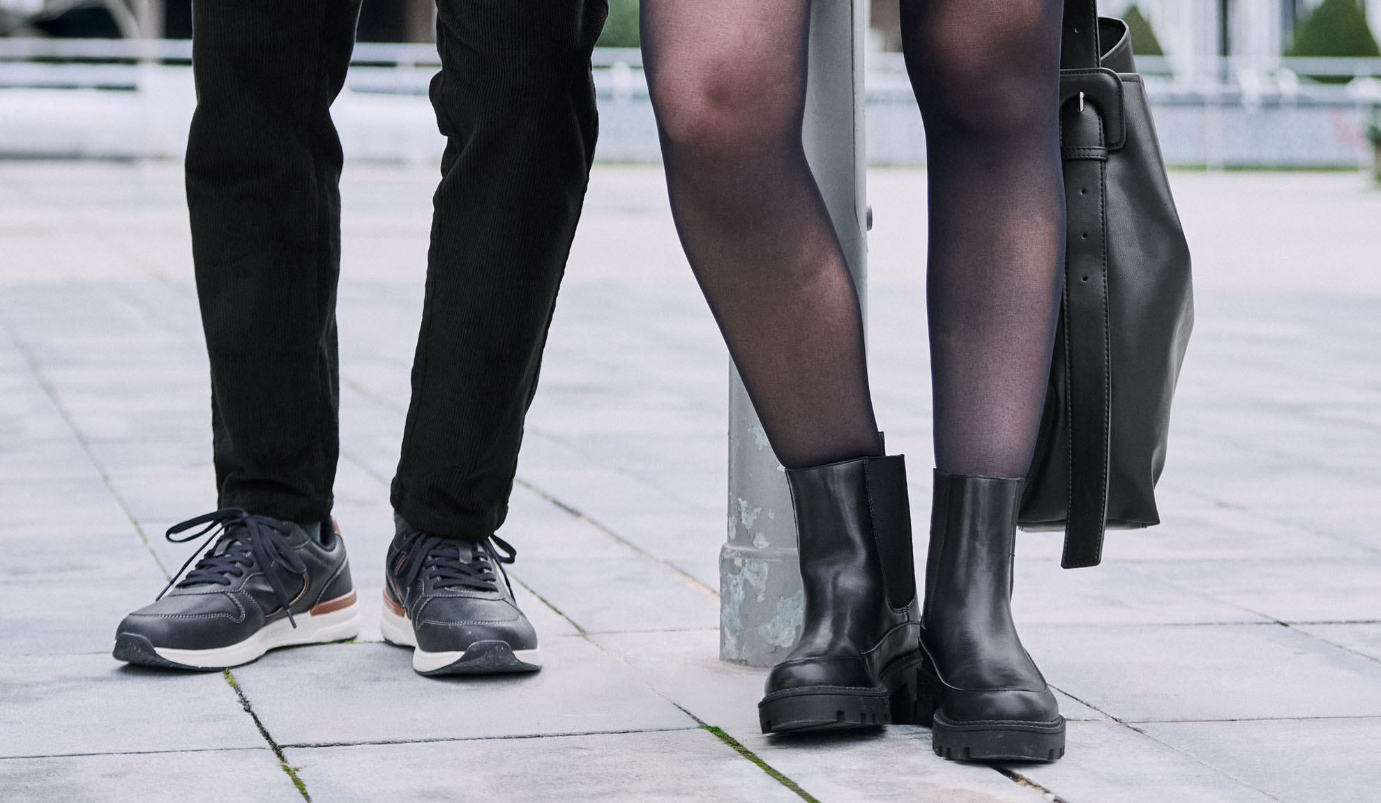 Gambe di due persone, una con pantaloni e sneakers, l'altra con collant, stivali e una borsa.
