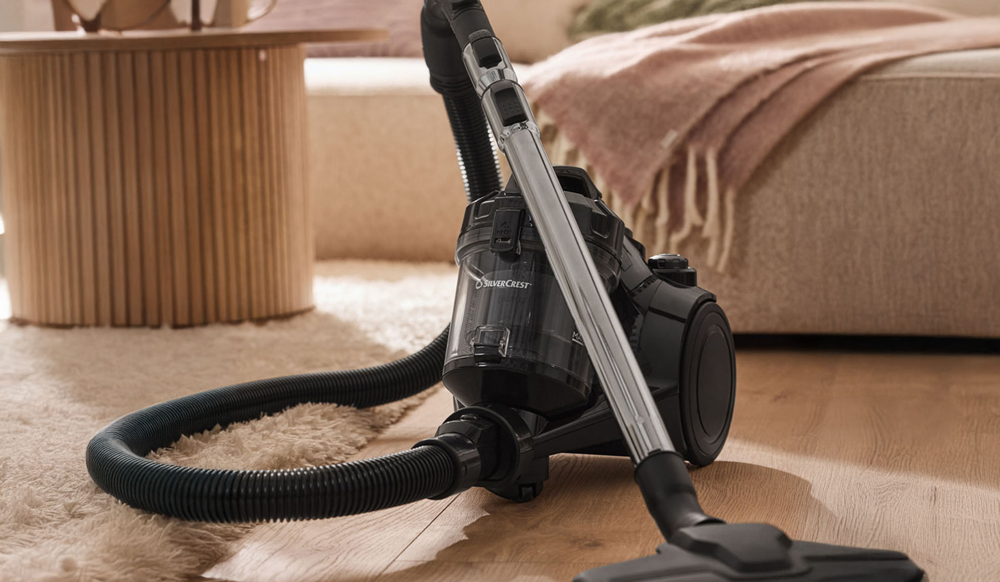 Aspirapolvere nero su pavimento in legno, con tappeto soffice e mobili sullo sfondo.