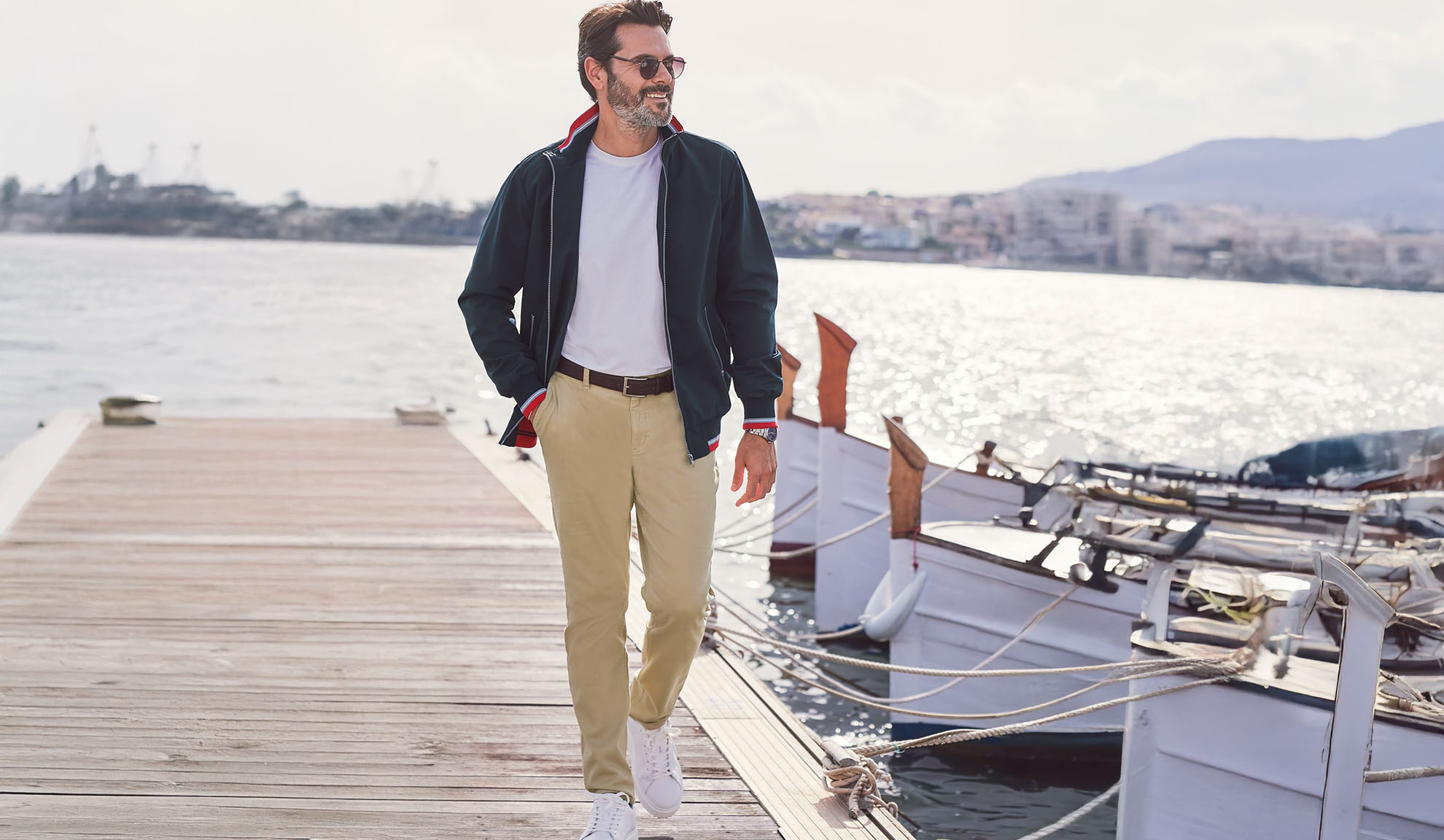 Mann in marineblauer Jacke, weißem T-Shirt und Khakihose auf einem Pier.