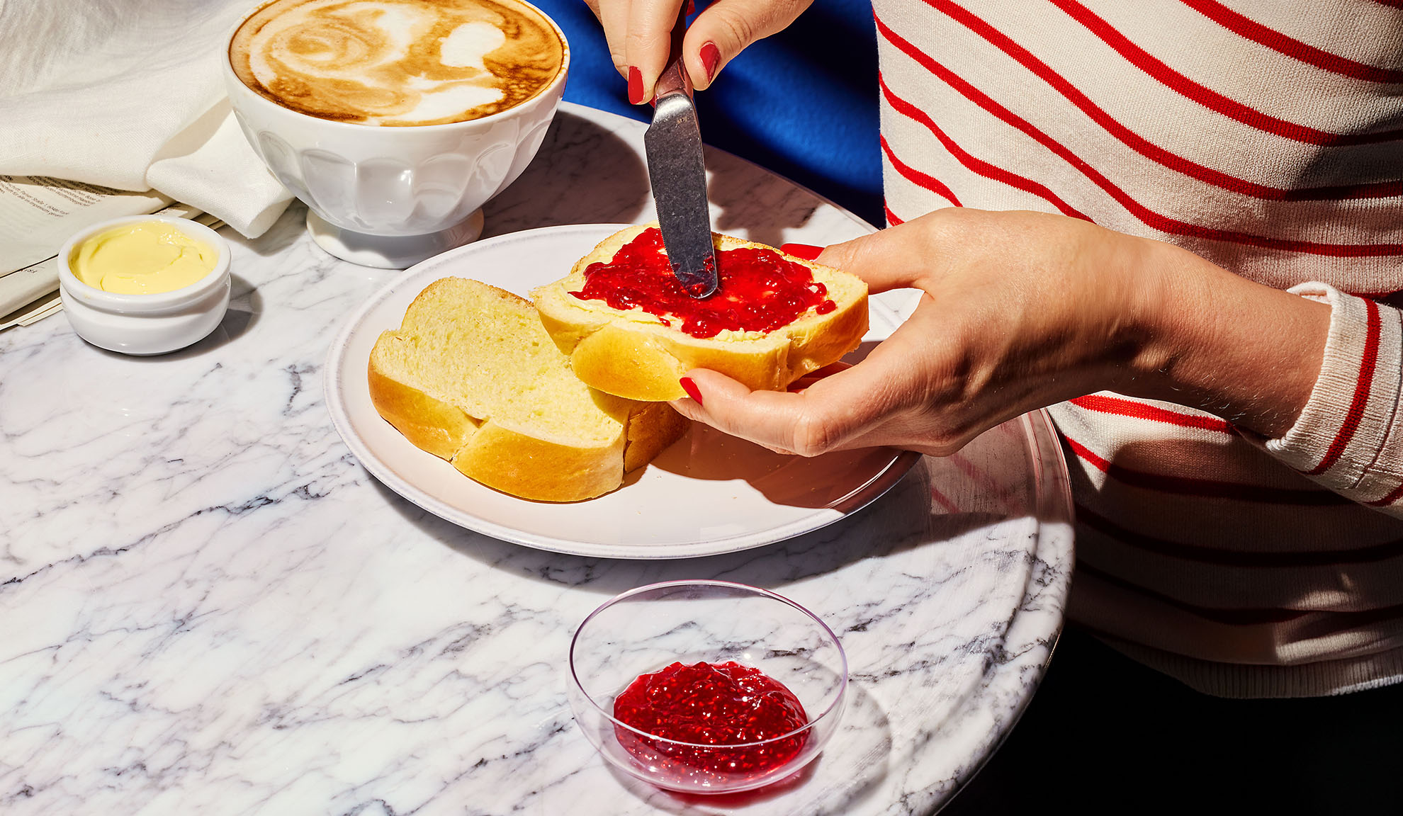 Hände streichen Marmelade auf Briochebrot, mit Kaffee und Butter auf Marmortisch.