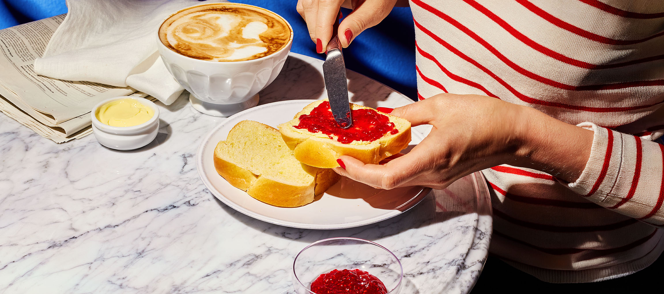 Hände streichen Marmelade auf Briochebrot, mit Kaffee und Butter auf Marmortisch.