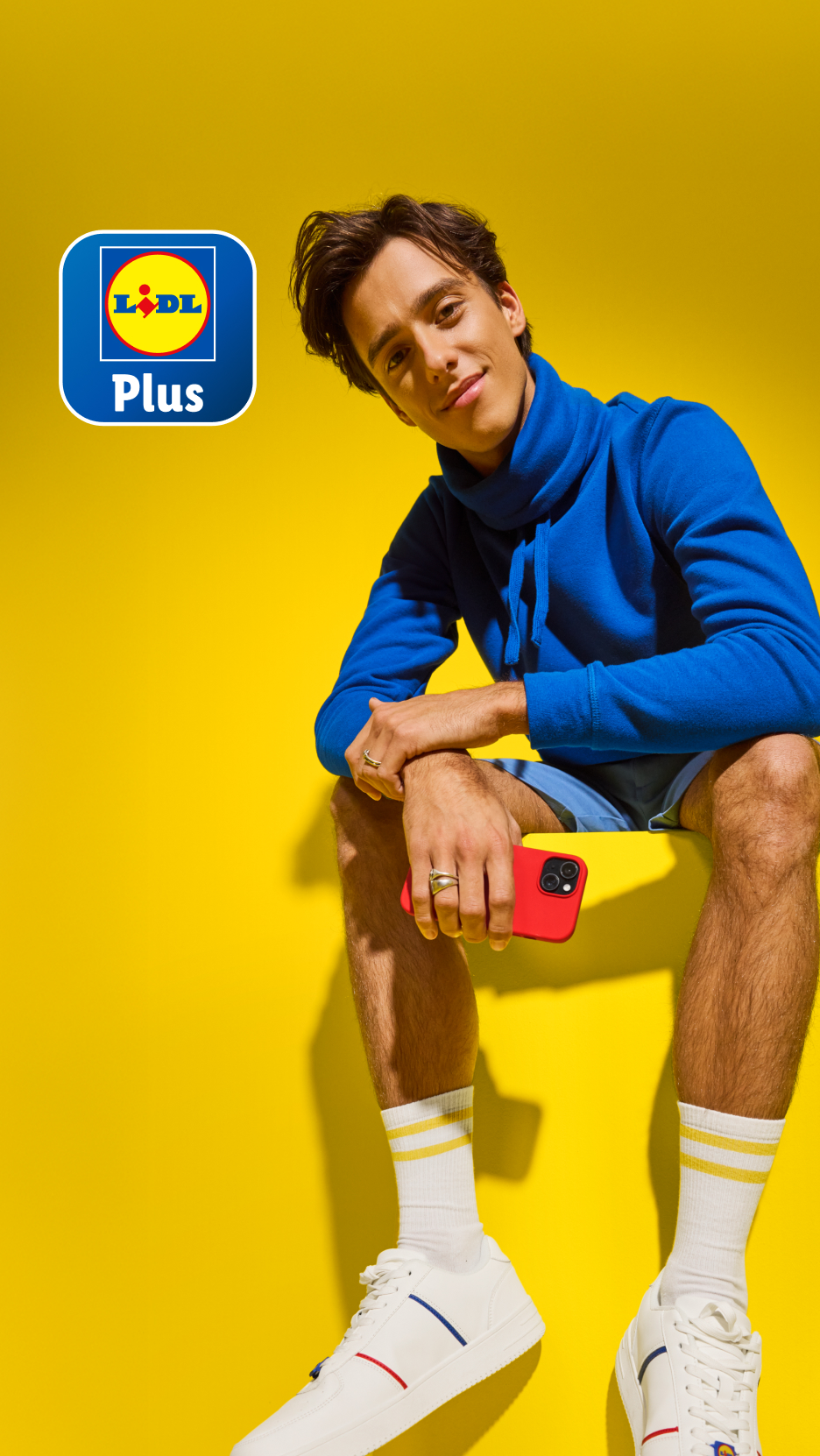 Lächelnder Mann in blauem Hoodie mit rotem Telefon, sitzt auf gelbem Hintergrund mit „Plus“-Logo.