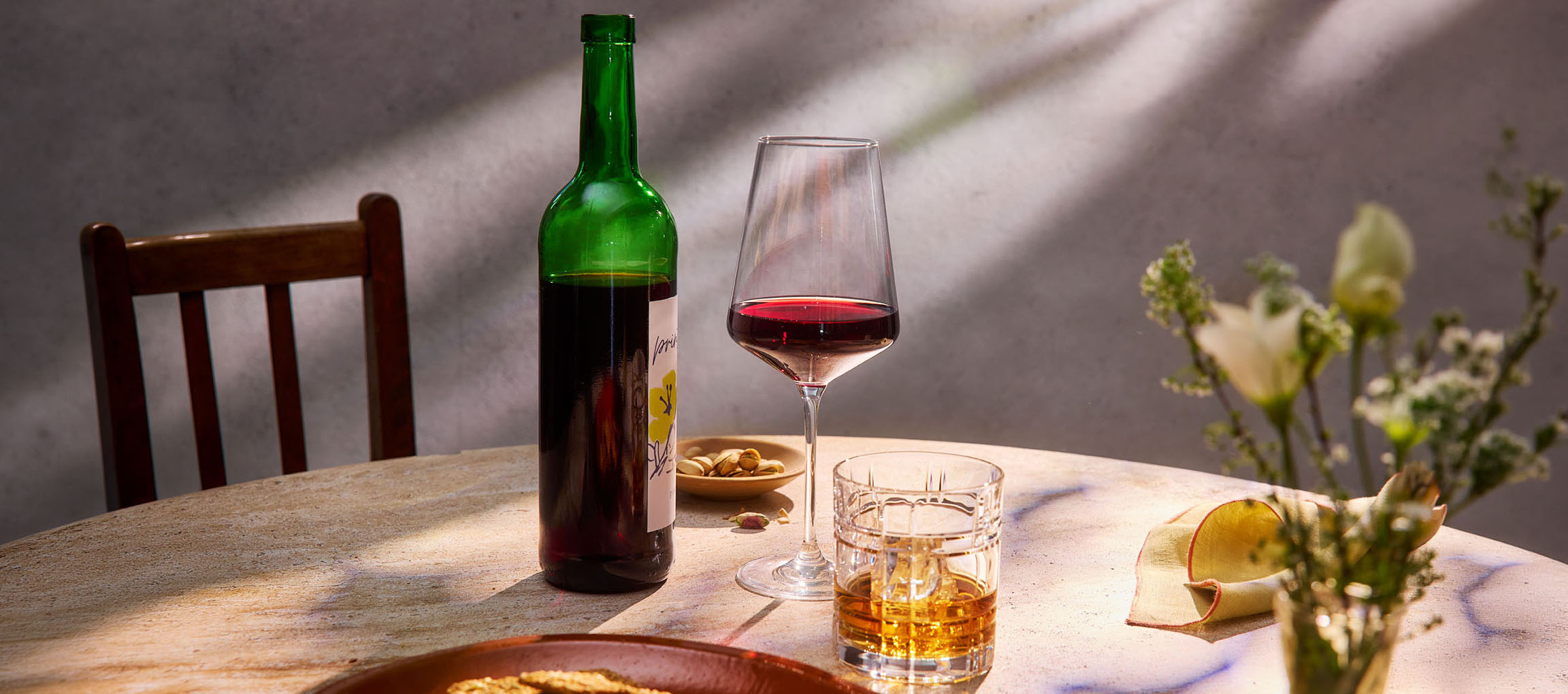 Flasche Rotwein, Weinglas und Whisky auf einem Tisch, mit Stuhl und Blumen im Hintergrund.