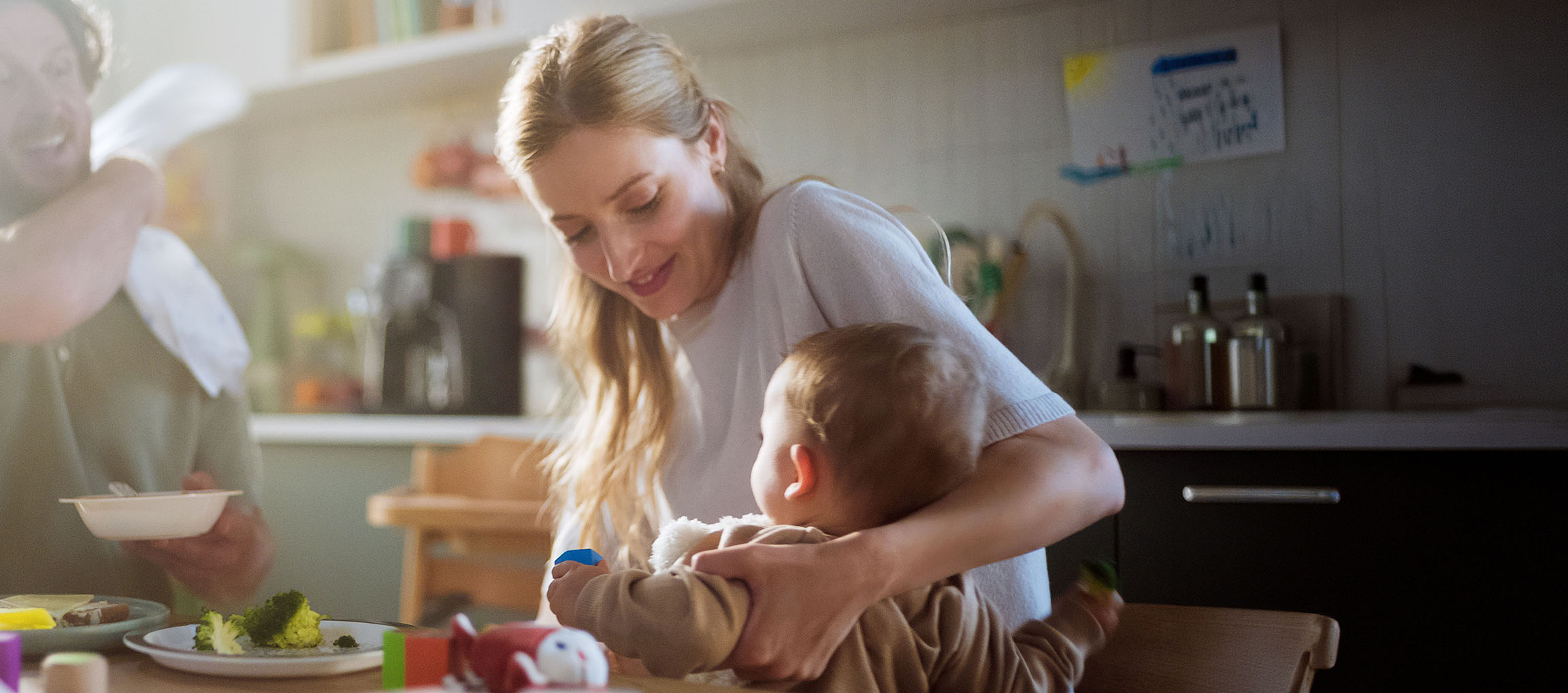Famiglia in cucina, madre che nutre il bambino, padre che mangia broccoli dal piatto.