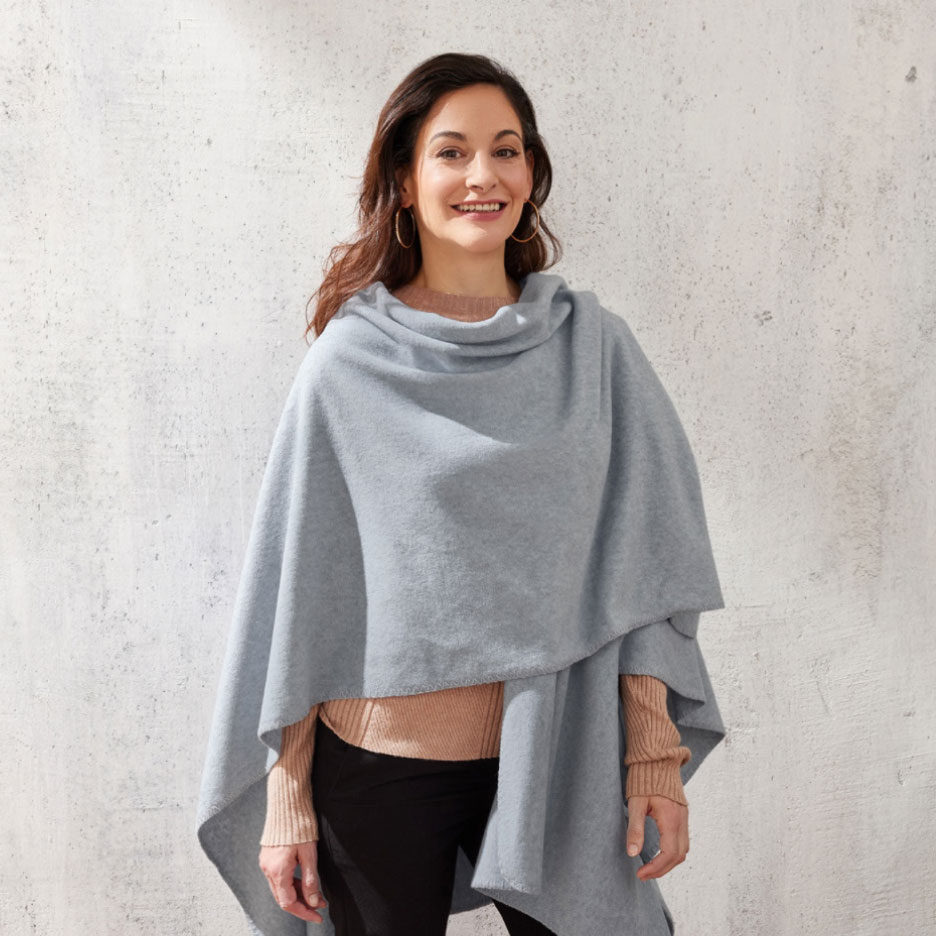 Lächelnde Frau trägt einen hellblauen Poncho über einem kamelfarbenen Pullover und schwarzen Hosen.