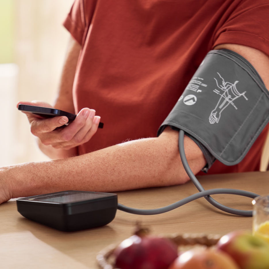Person benutzt Blutdruckmessgerät und Smartphone an einem Tisch mit Obst.