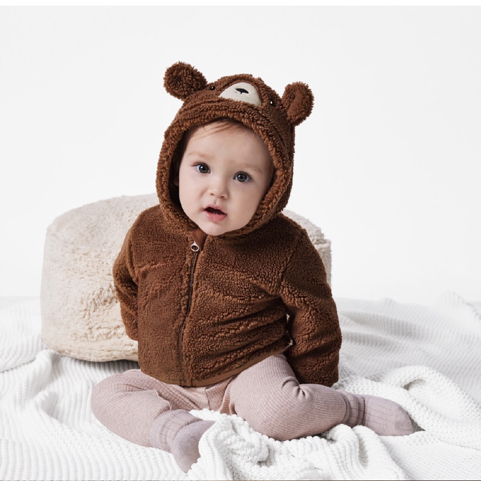 Baby in brauner Fleecejacke mit Bärenkapuze und rosa Leggings auf einer weißen Decke.