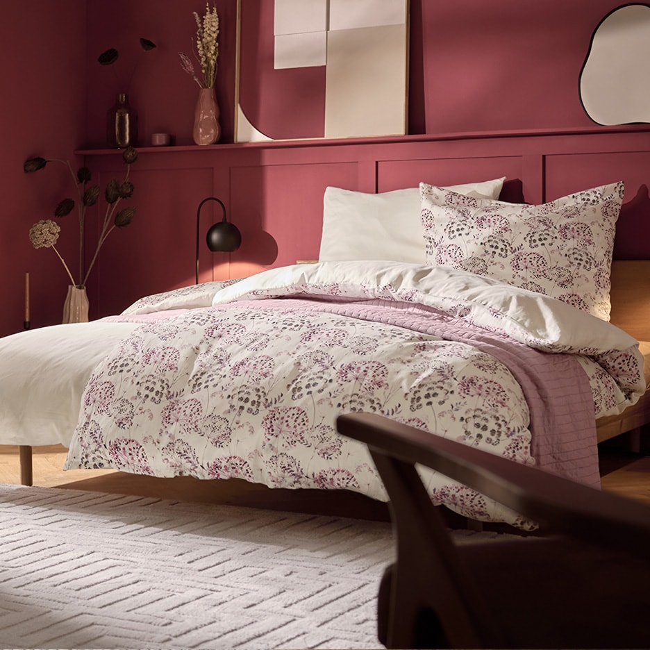Schlafzimmer mit himbeerfarbenen Wänden, Bett mit geblümter Bettdecke und weißen Kissen.
