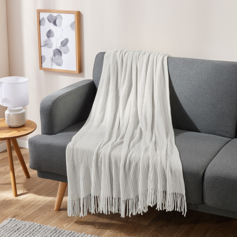 Ein weißer Überwurf mit Fransen, drapiert über ein graues Sofa in einem modernen Wohnzimmer.
