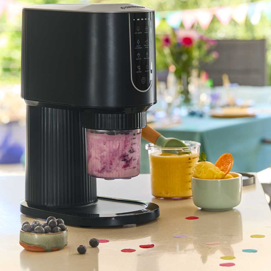 Schwarze Eismaschine mit lila Eis, umgeben von Mango-Eis und Blaubeeren.