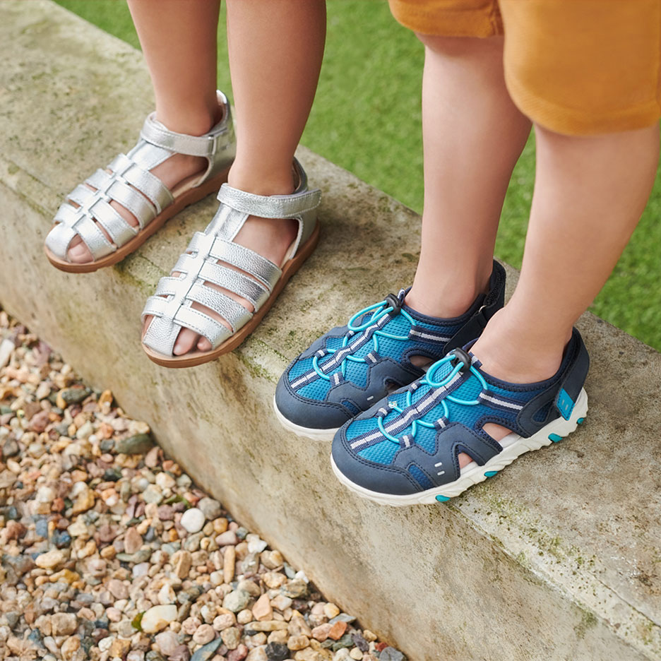 Deux paires de sandales pour enfants : argentées et bleues, sur un trottoir.