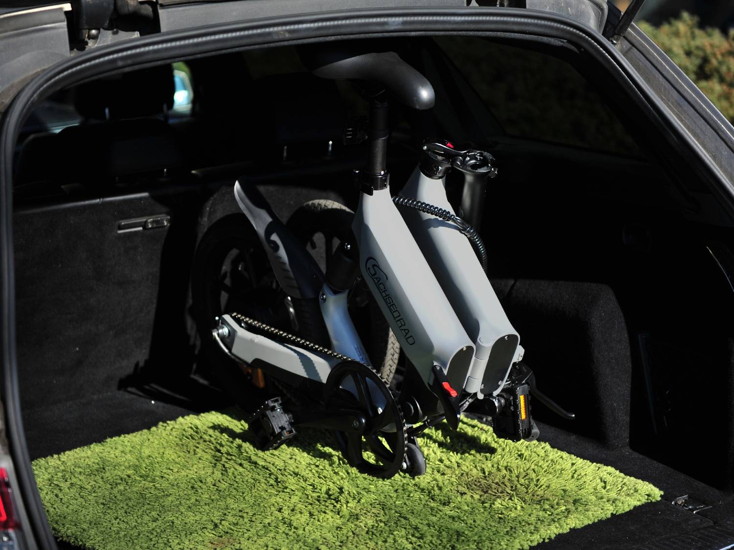 Bicicletta elettrica pieghevole grigia riposta nel bagagliaio di un'auto su un tappeto verde.