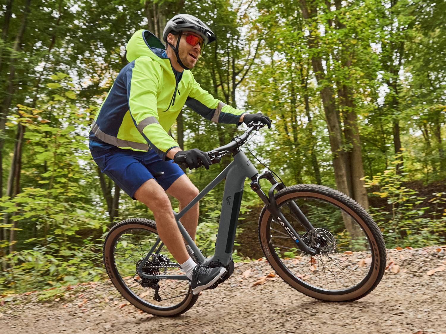 Uomo in bici elettrica su sentiero nel bosco, indossa giacca a vento gialla e casco.