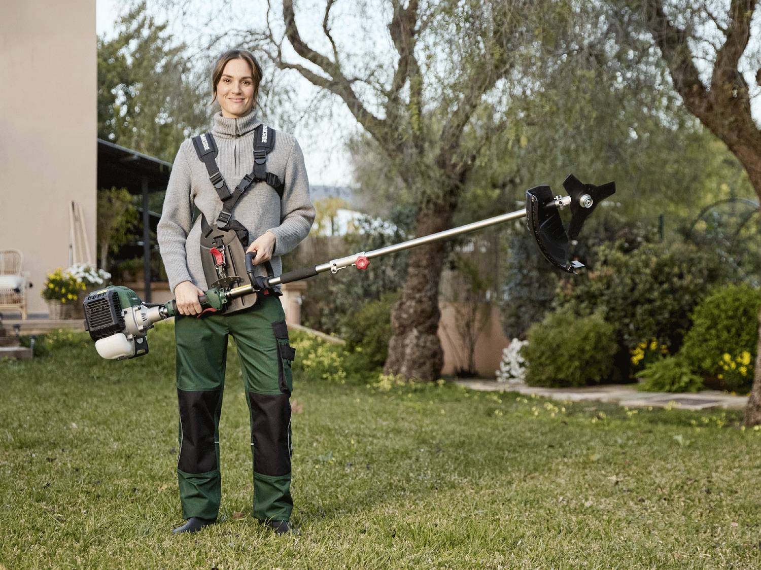 Donna sorridente con decespugliatore a benzina e imbracatura in un giardino verde.