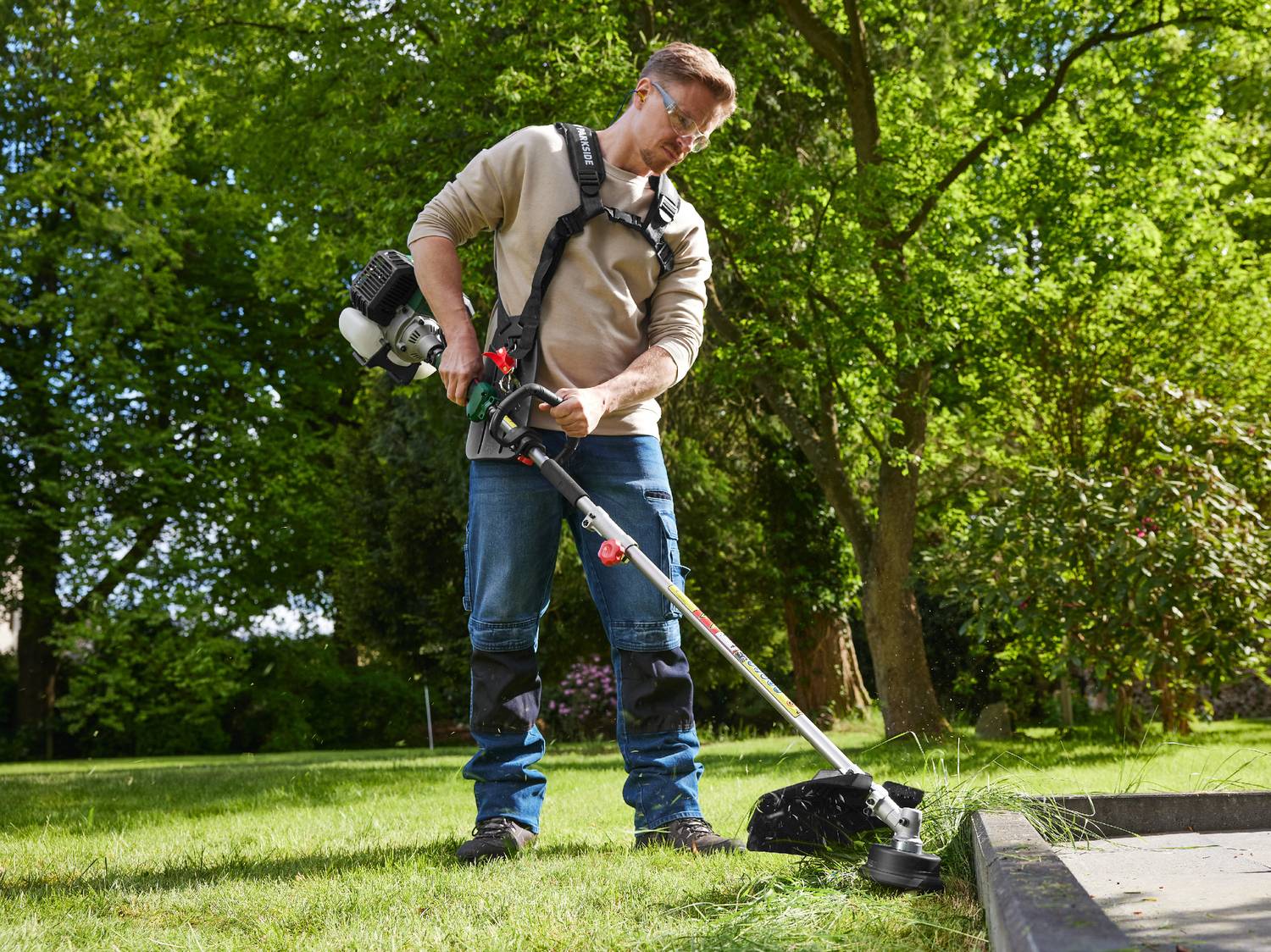 Uomo che usa un decespugliatore a benzina con imbracatura in un giardino verde.