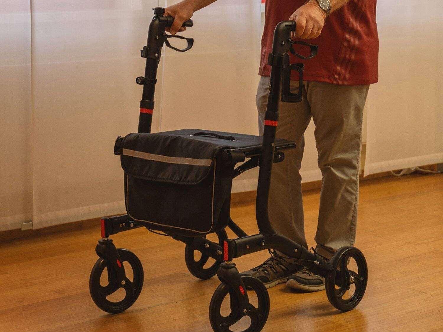 Eine Person steht mit einem schwarzen Rollator mit Aufbewahrungstasche und vier Rädern.