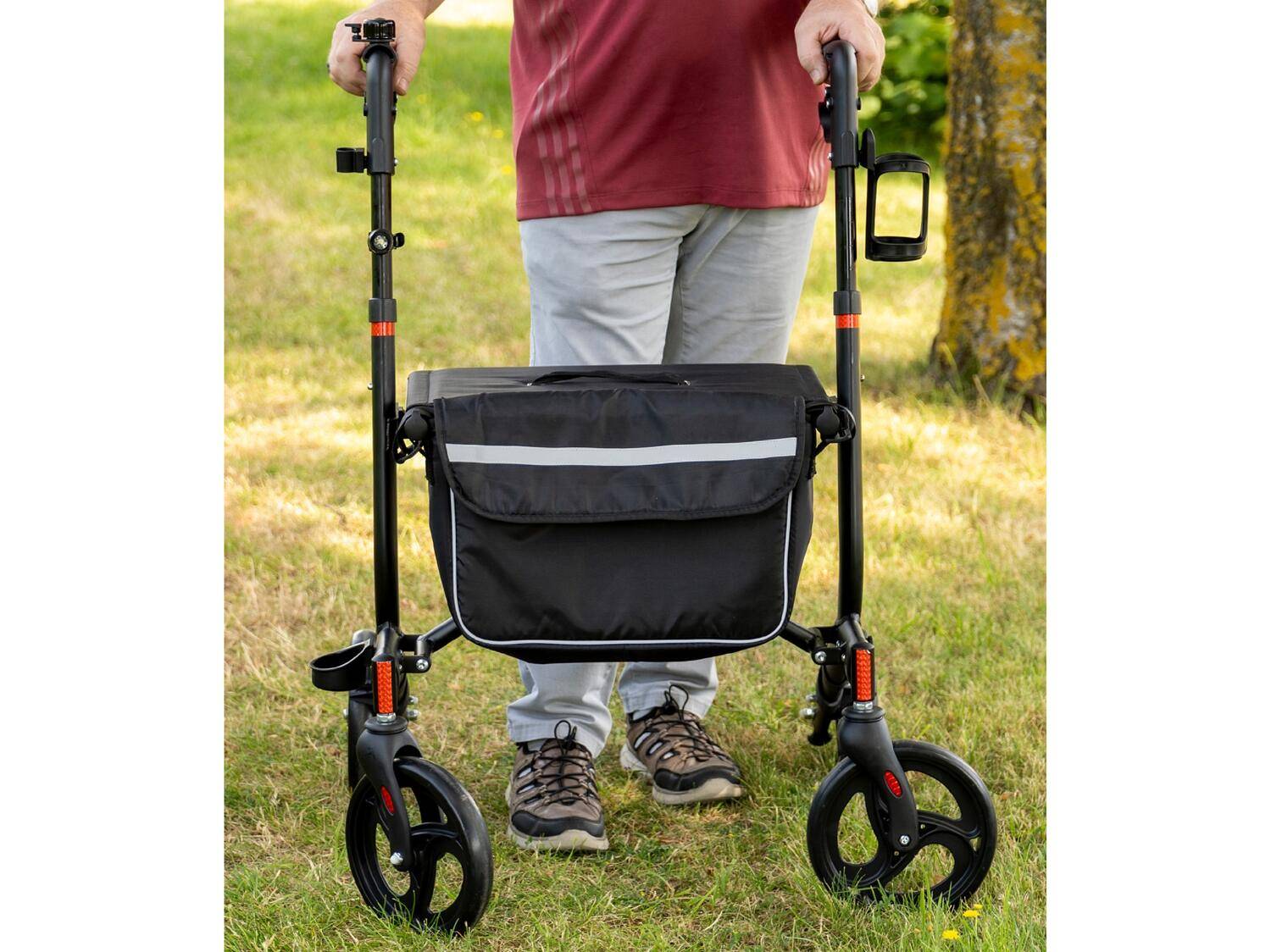 Eine Person benutzt einen schwarzen Rollator mit Tasche und Getränkehalter auf einer Wiese.