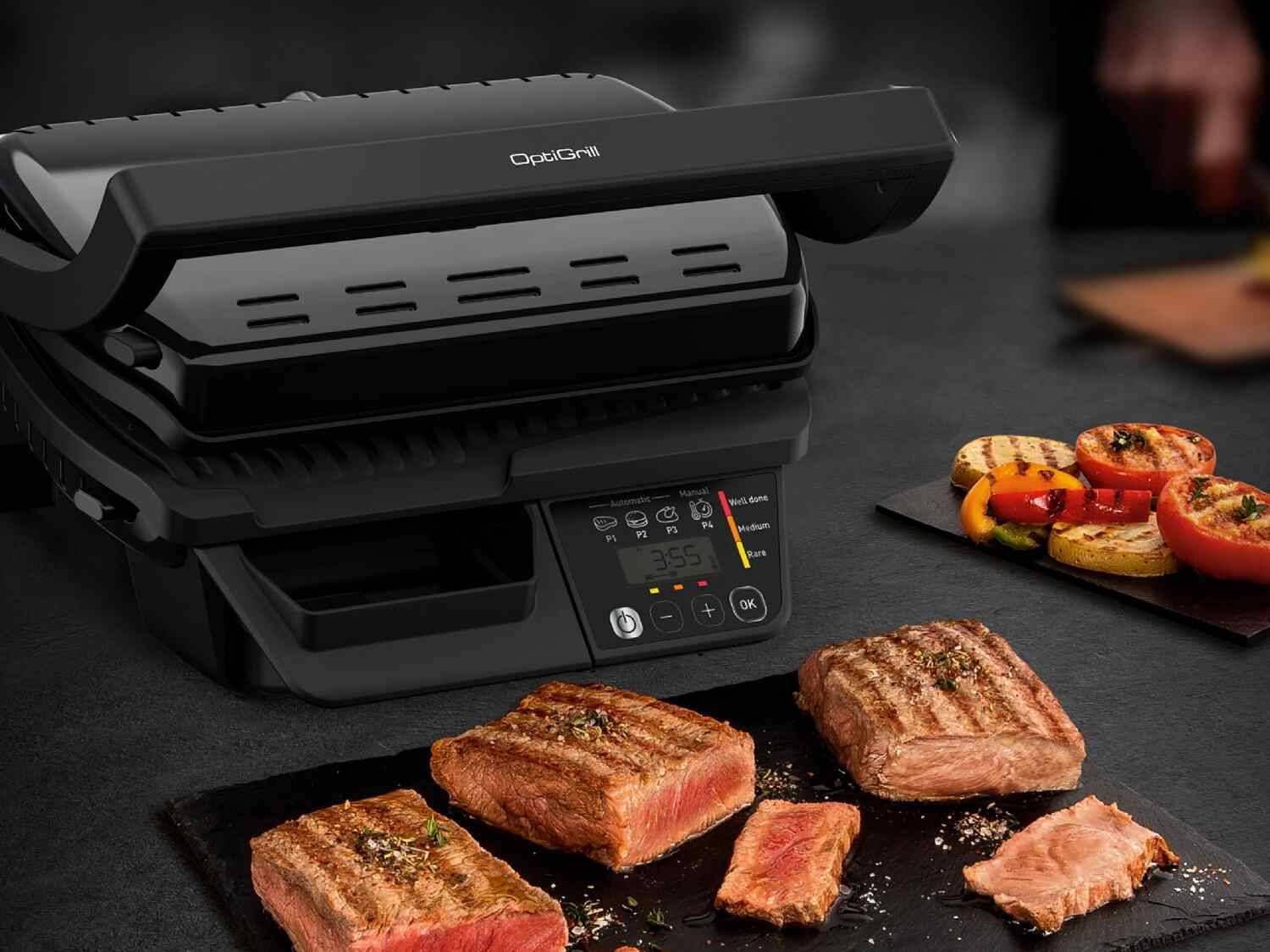 OptiGrill-Grill mit Steaks und gegrilltem Gemüse auf dunklem Untergrund.