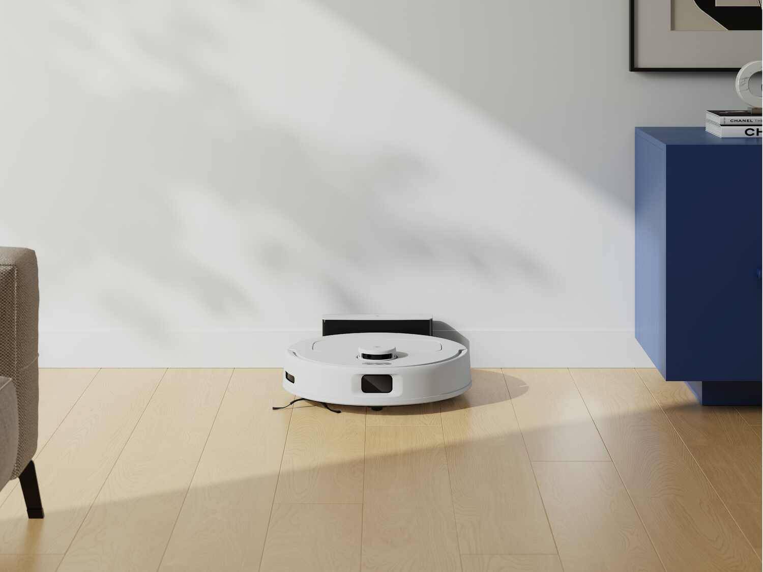 Ein weißer Saugroboter auf hellem Holzboden, vor einer weißen Wand und einem blauen Schrank.