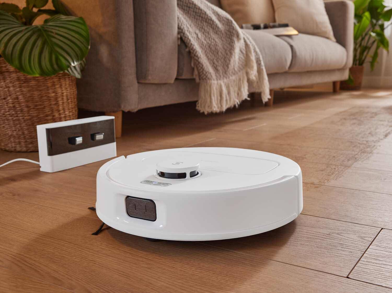 Weißer Saugroboter auf Holzboden mit Ladestation und Sofa.