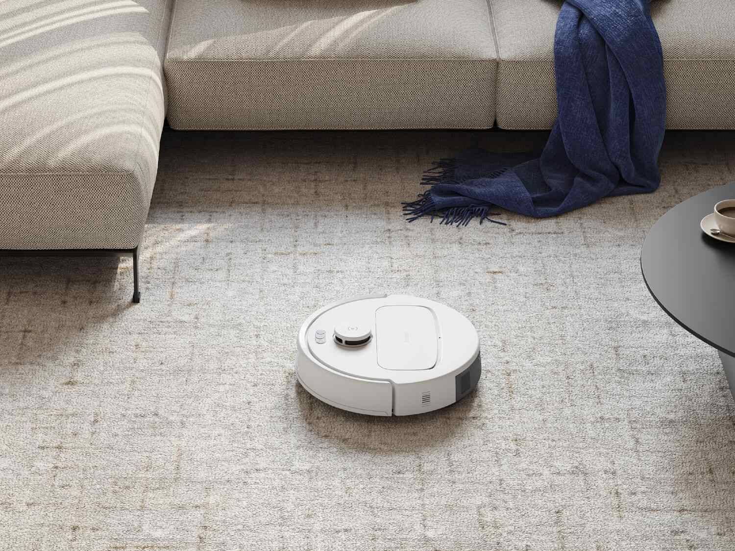 Ein weißer Saugroboter auf einem Teppich, neben einem Sofa und einem Couchtisch.