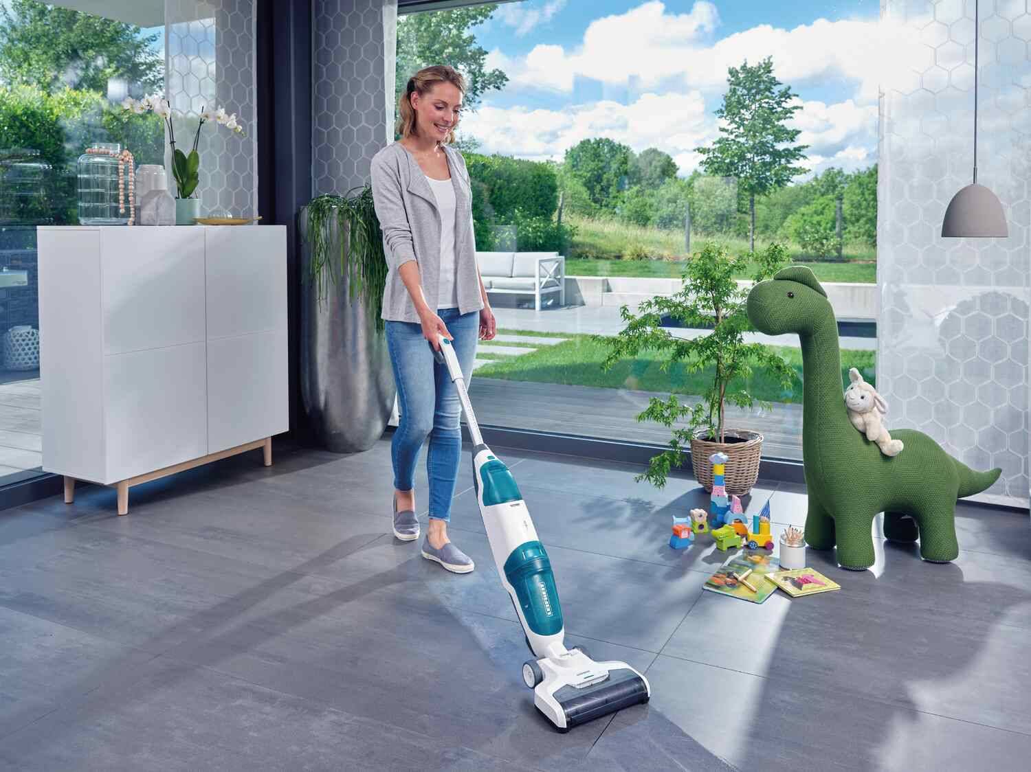Frau reinigt den Boden mit einem kabellosen Staubsauger, mit einem Spielzeugdinosaurier und bunten Bausteinen.