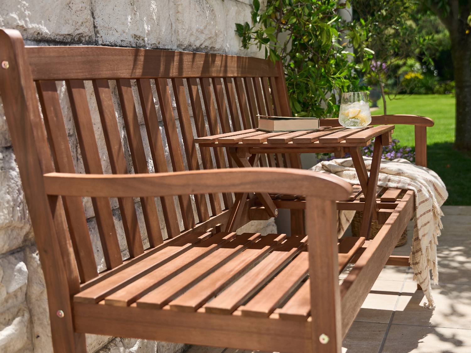 Panca da giardino in legno con tavolo integrato, libro e bevanda rinfrescante.