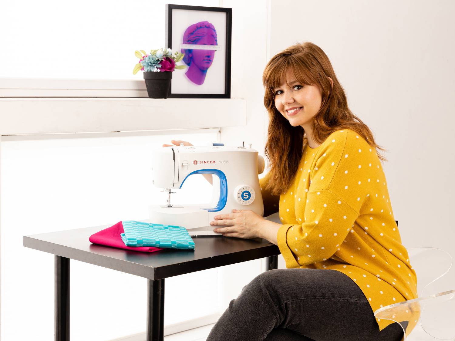 Femme souriante assise à une table avec une machine à coudre et des tissus.