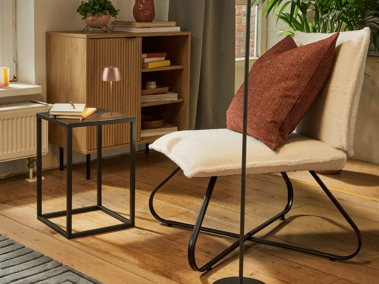 Ein gemütliches Wohnzimmer mit einem cremefarbenen Bouclé-Loungesessel, einem schwarzen Metall-Beistelltisch und einem Holzschrank.