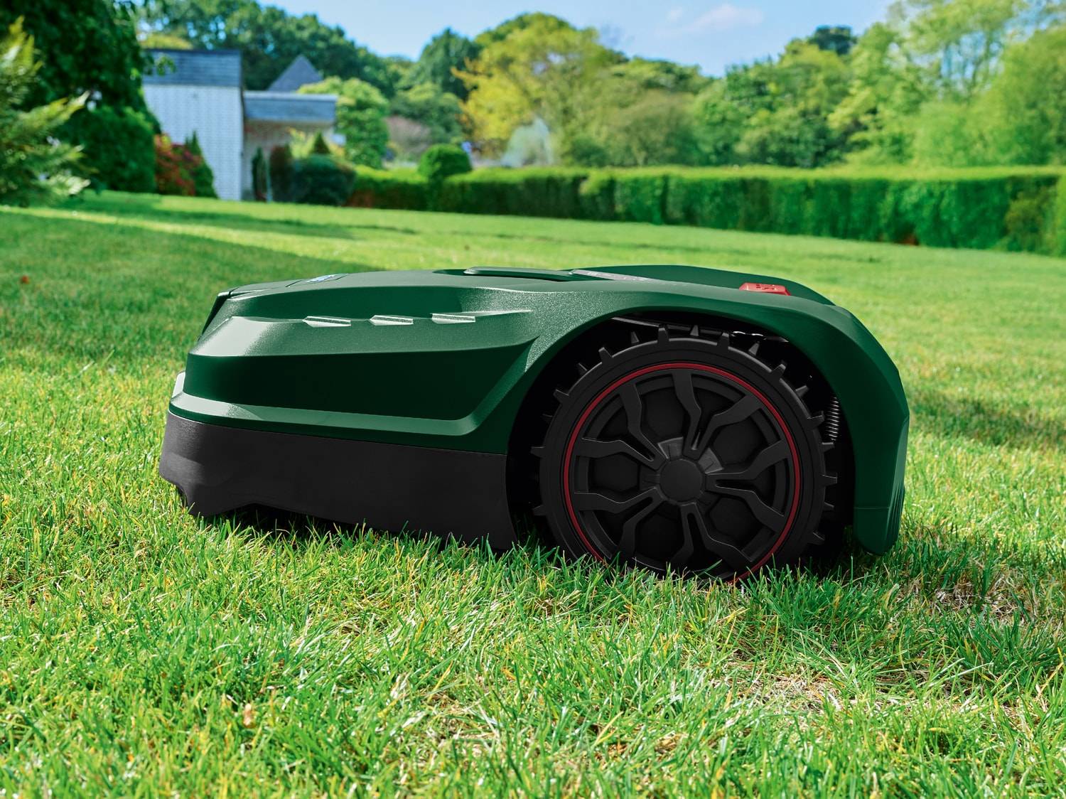 Robot tondeuse vert sur pelouse verte avec maison et arbres en arrière-plan.