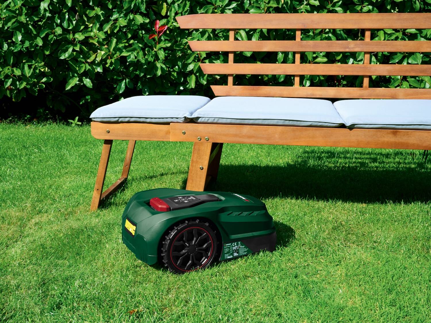 Robot tondeuse vert sur pelouse verte avec banc en bois et coussins clairs.