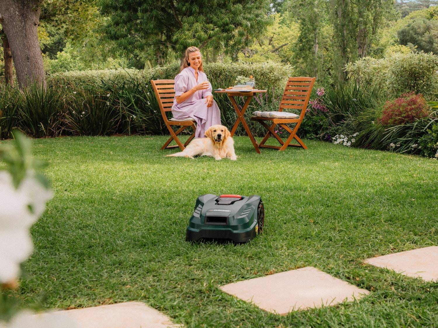 Une tondeuse robot sur la pelouse, avec une femme et un chien assis à une table de jardin en arrière-plan.