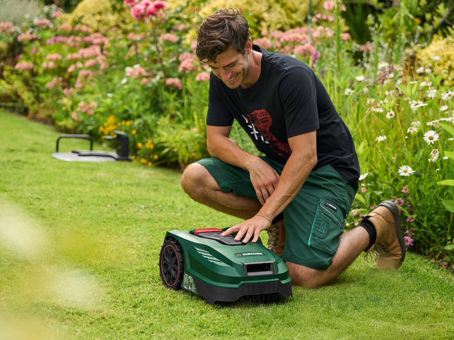 Homme souriant accroupi sur l'herbe, réglant une tondeuse robot verte dans un jardin fleuri.