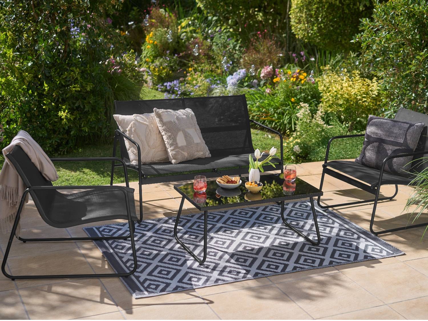 Salon de jardin noir avec canapé, fauteuils, table basse et tapis géométrique sur terrasse.