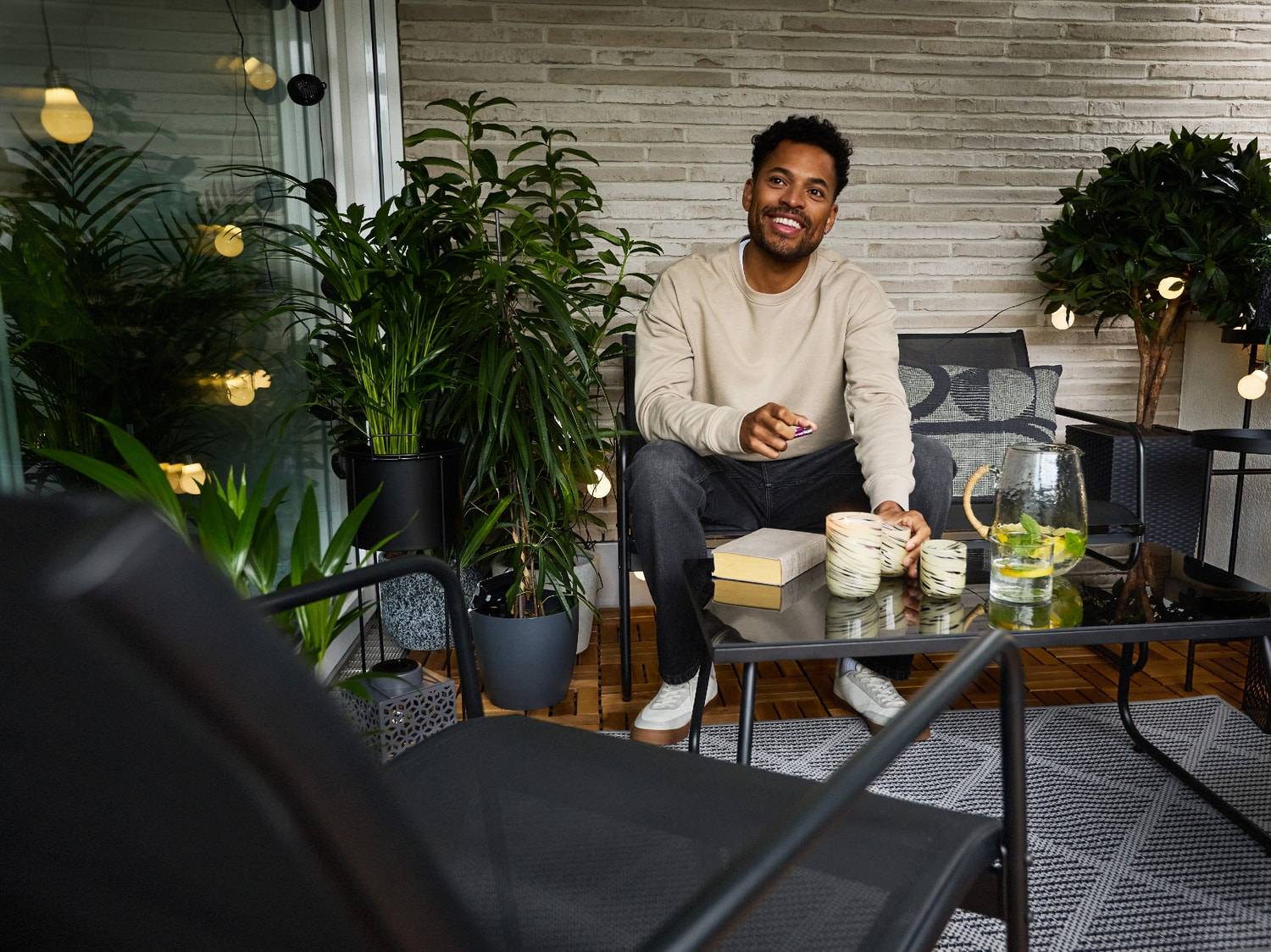 Homme souriant assis sur un balcon aménagé avec des meubles d'extérieur, des plantes et des lumières décoratives.