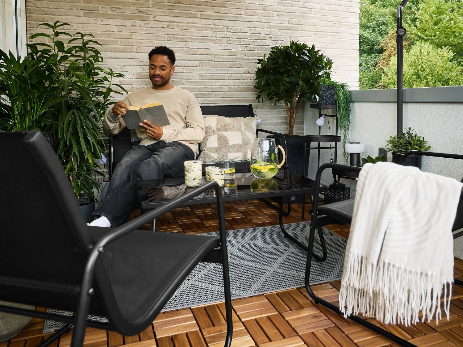 Homme lisant sur un canapé de balcon avec table, chaises et plantes.