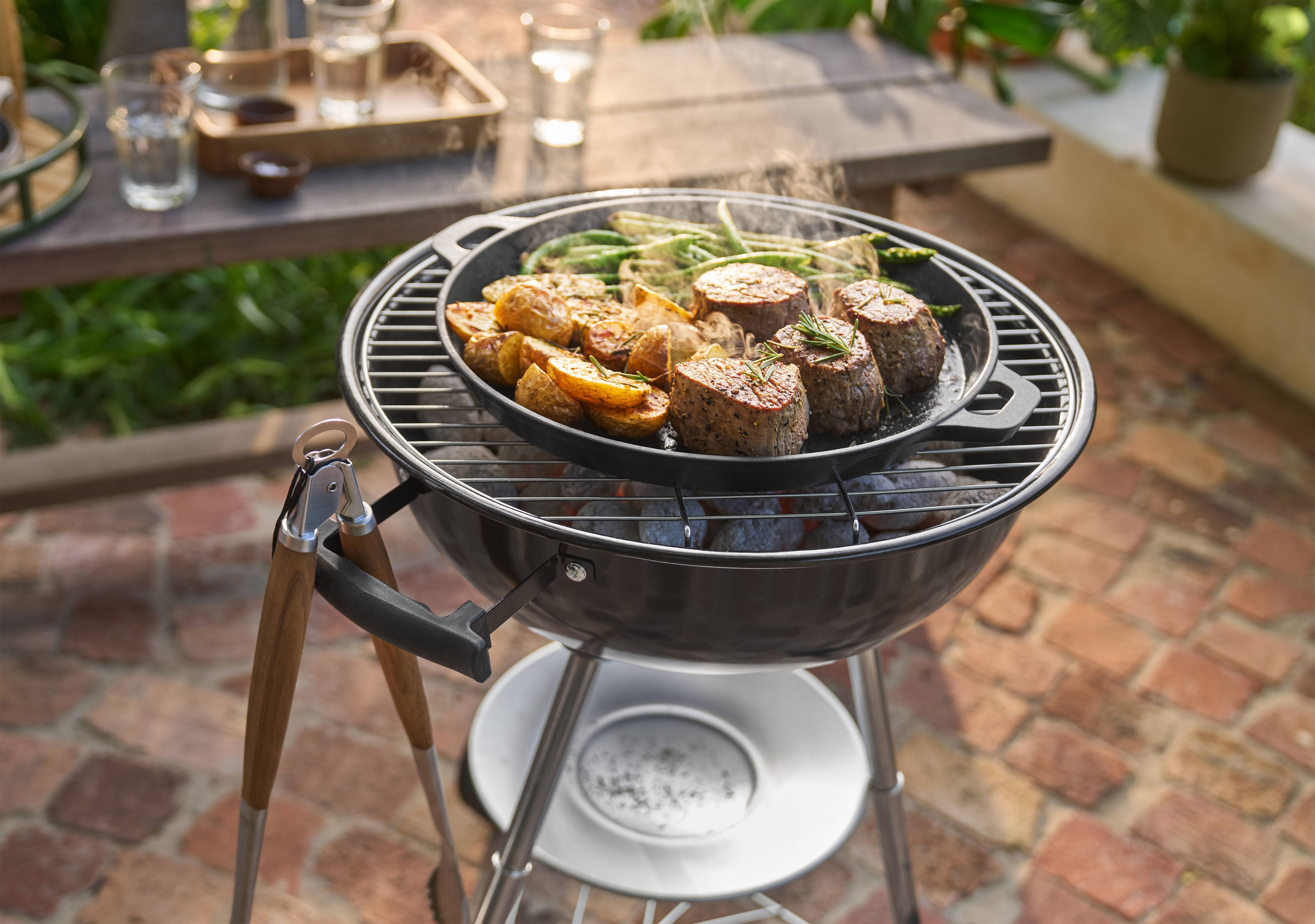 Barbecue avec viande, pommes de terre et haricots verts cuisant dans une poêle en extérieur.