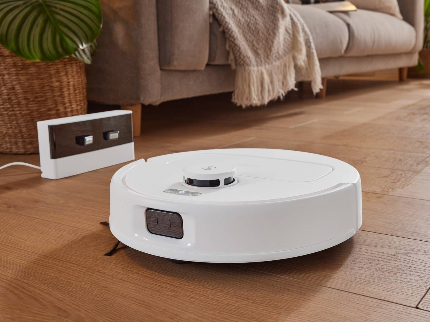 Un robot aspirateur blanc est posé sur un parquet en bois à côté de sa station de charge.