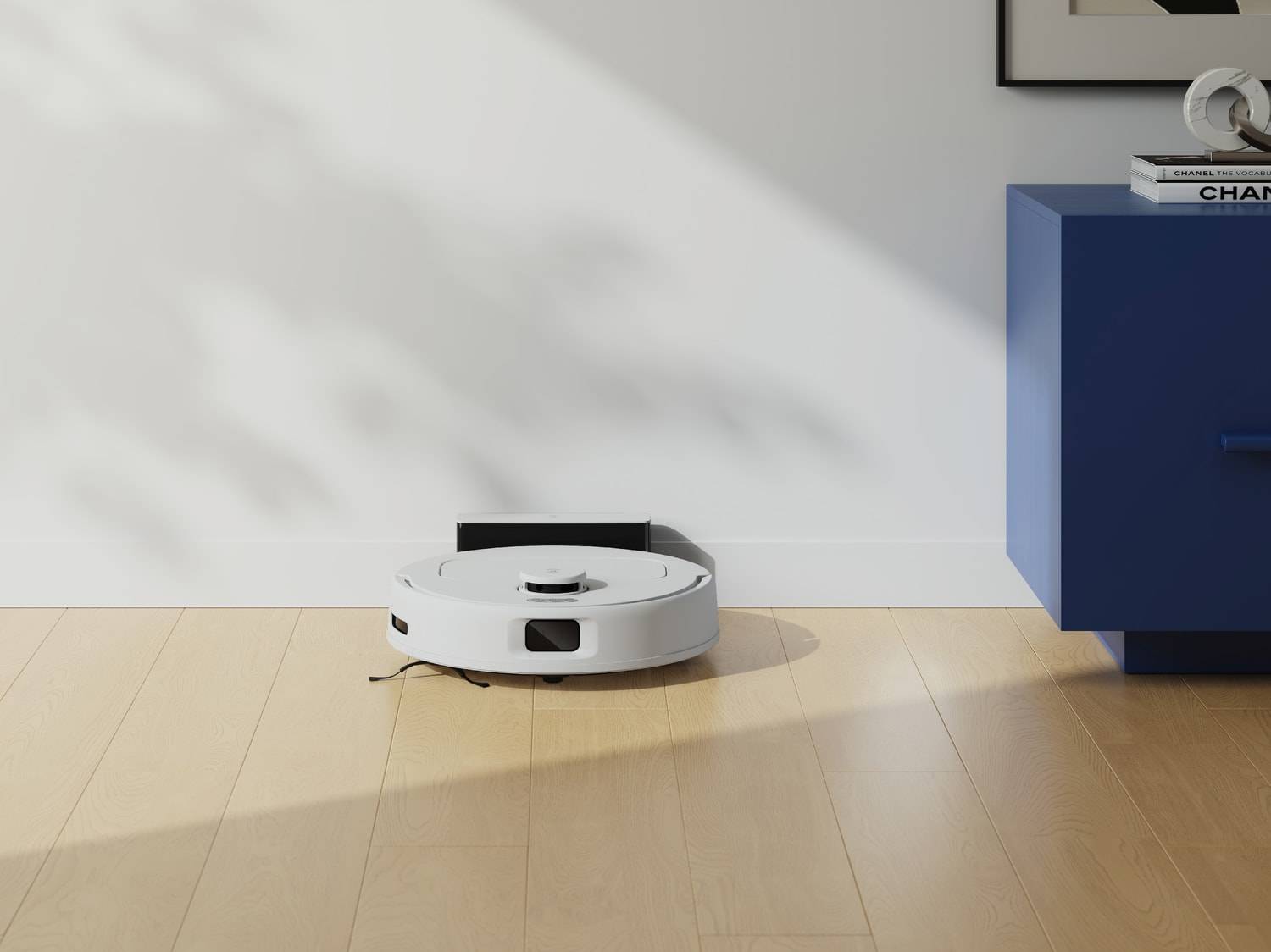 Un robot aspirateur blanc sur un parquet en bois, devant un mur blanc et un meuble bleu.